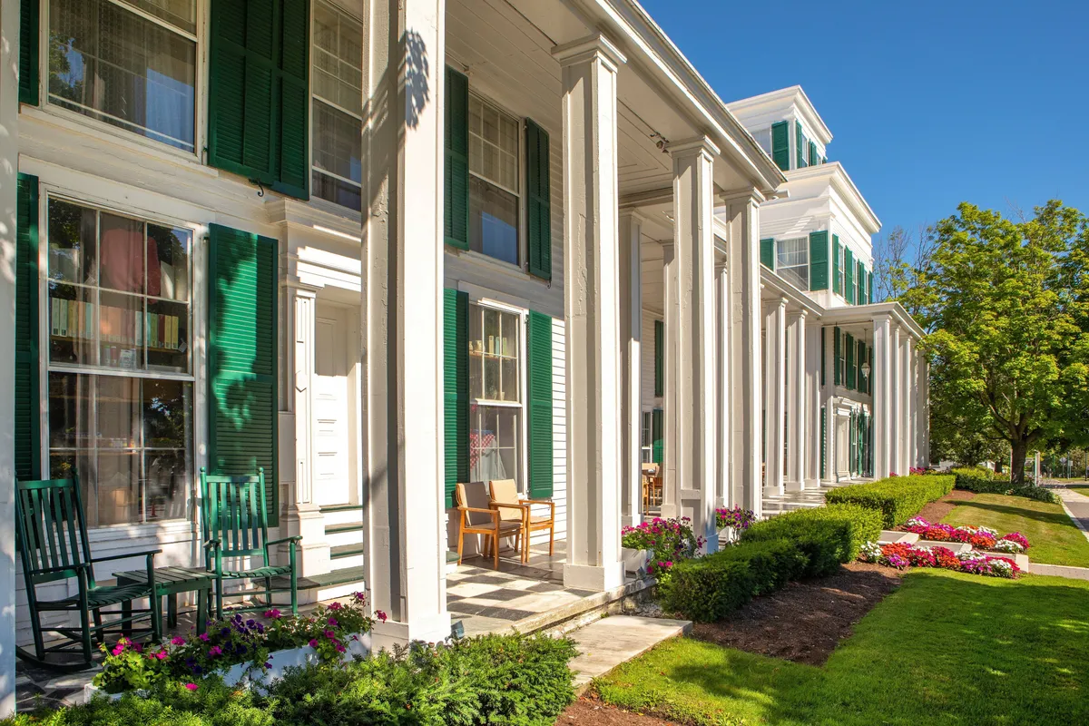 This image shows the stately exterior of The Equinox, featuring classic New England architecture with white columns, inviting green shutters, and an expansive porch lined with rocking chairs. Flower beds add vibrant color to the scene, and the overall mood is peaceful and welcoming—ideal for couples seeking a romantic, relaxing escape. The porch offers views of the gardens and hints at the social, convivial atmosphere within.