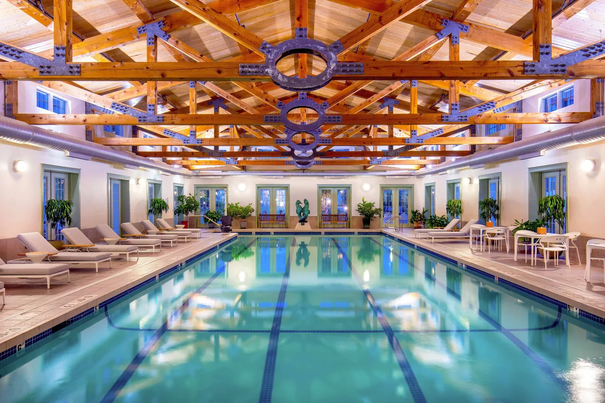 An elegant indoor swimming pool space with warm wooden beams, abundant lounge chairs, lush green plants, and floor-to-ceiling windows that invite natural light. The tranquil ambiance makes it ideal for swimming laps or relaxing after a spa session.