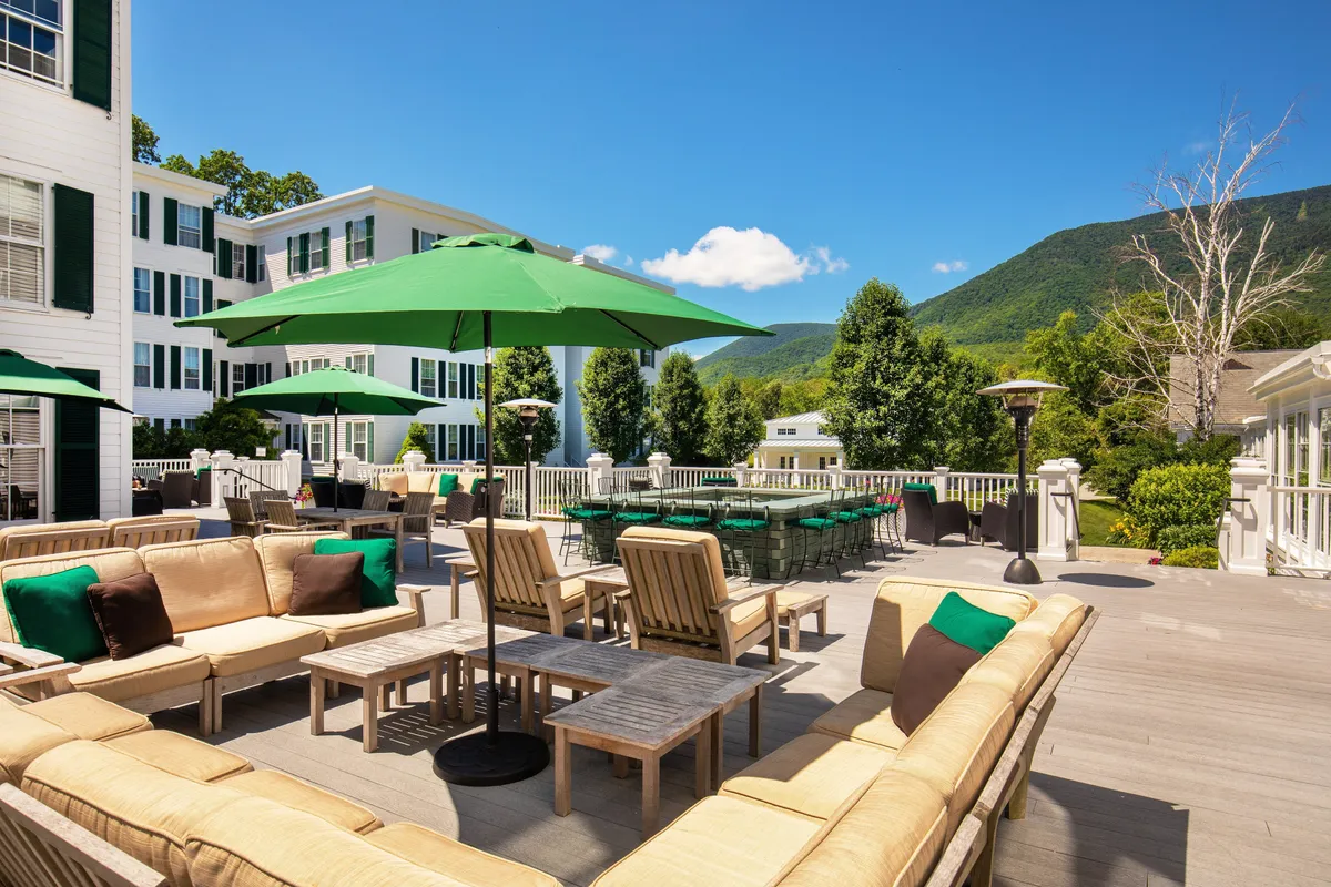 An inviting outdoor dining area shaded by green umbrellas, with sectional lounge seating overlooking breathtaking mountain views. Guests relax, socialize, and enjoy meals or drinks as the sun sets, blending indoor comfort with the natural beauty just outside.