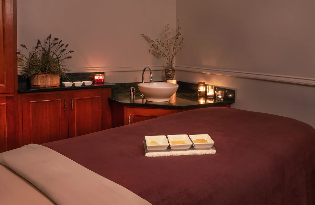 A serene spa treatment room featuring a comfortable massage table, a bowl of aromatherapy oils, softly glowing candles, and a decorative plant. The calming decor provides an immediate sense of relaxation and luxurious care.
