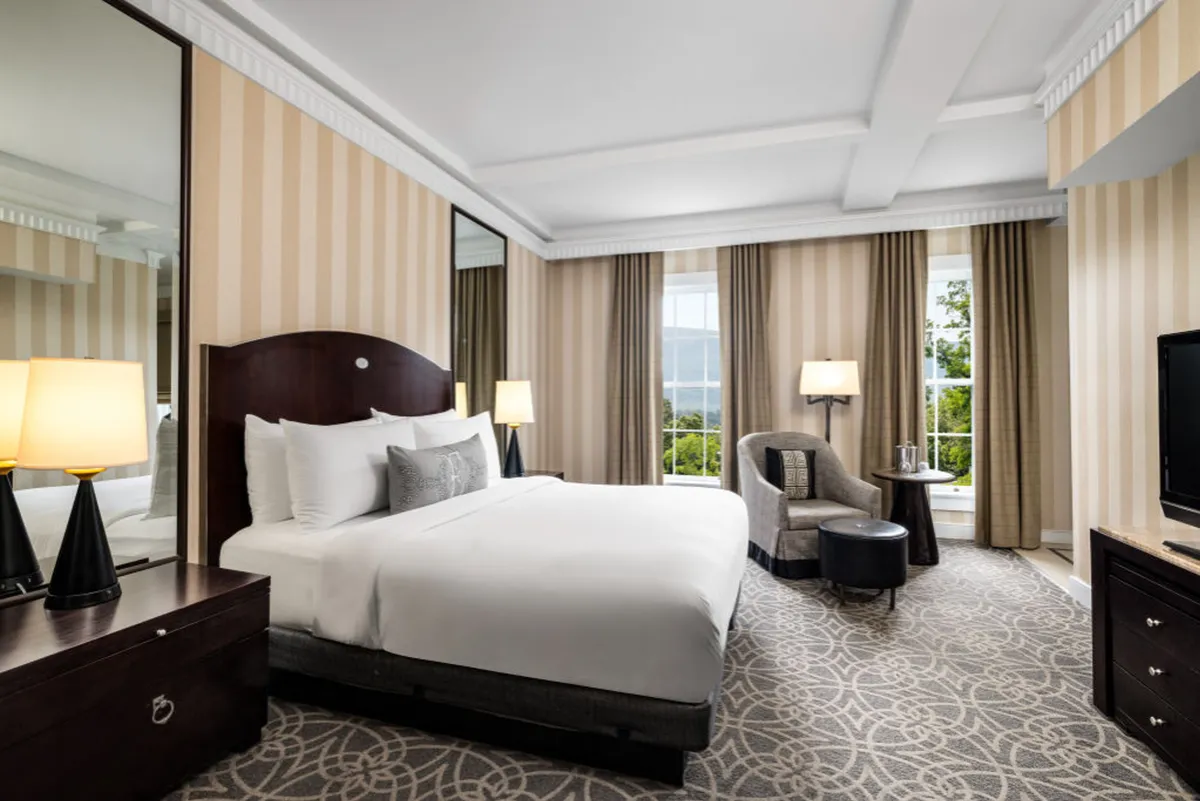 The image showcases a beautifully appointed studio interior at The Equinox Golf Resort & Spa in Manchester, Vermont. The scene centers on a plush bed adorned with crisp linens, flanked by elegant lighting fixtures that cast a warm, tranquil glow. A comfortable chair sits invitingly by the window, perfect for reading or sipping morning coffee while natural light floods the space. A sleek TV is positioned for cozy evenings, and the overall atmosphere is serene and sophisticated, blending classic New England charm with modern comforts.