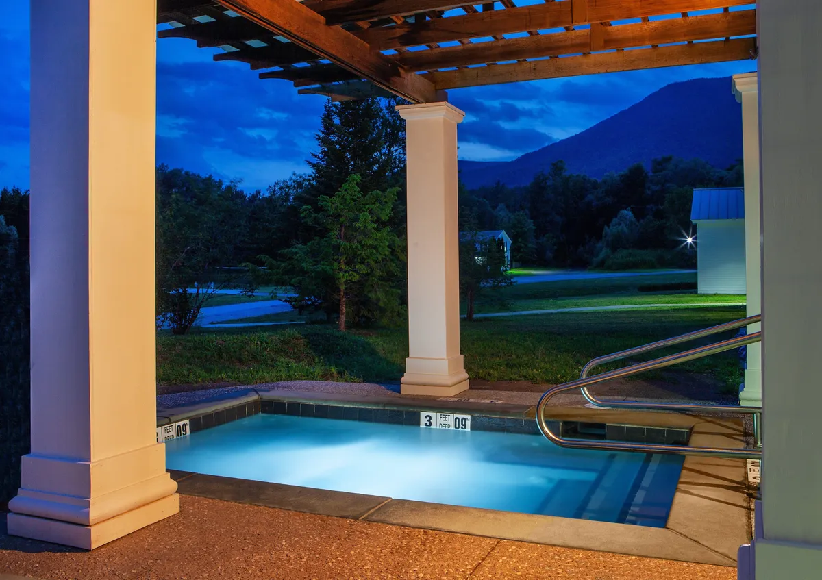 An inviting illuminated pool sits in the foreground, surrounded by a pergola structure, with a dramatic mountain backdrop under a serene evening sky. The peaceful ambiance suggests moments of relaxation after a day of adventure, while the proximity to nature is front and center.