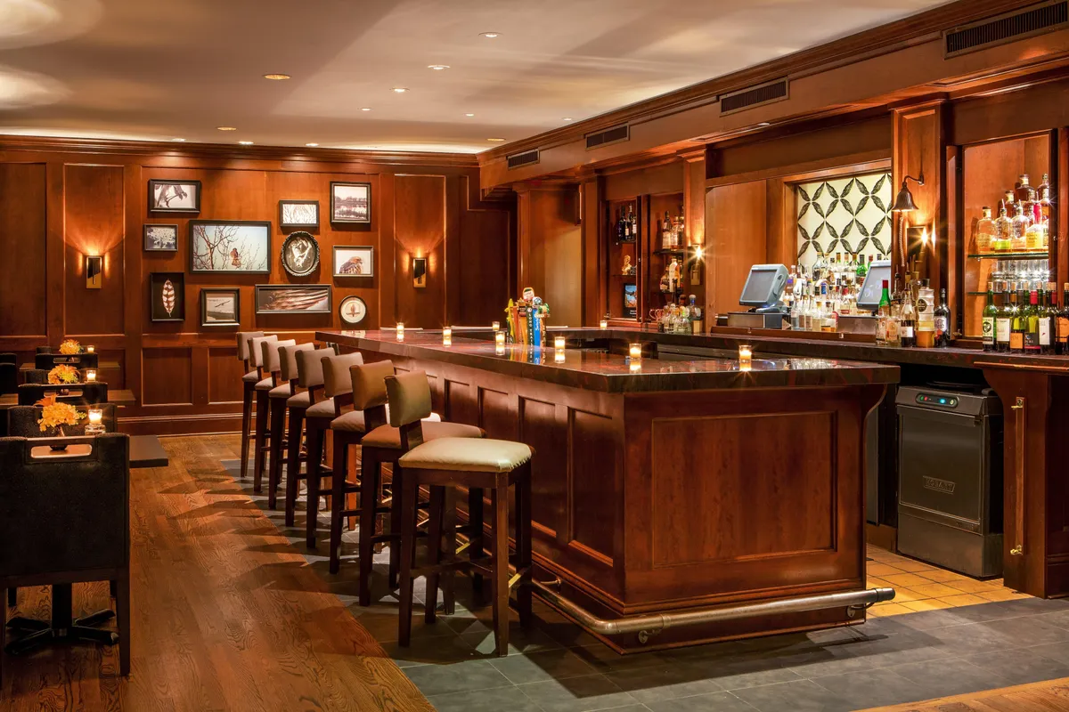 A sleek, cozy bar interior with a gleaming counter, plush bar stools, and shelves of premium spirits. The space is intimate and elegant, ideal for relaxed drinks, intimate conversations, or professional networking. Framed artwork provides a touch of local charm.